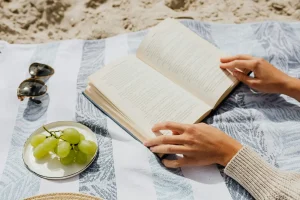 Persona leyendo un libro en la playa con uvas y gafas de sol sobre una toalla