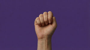 Mano levantada en forma de puño sobre un fondo morado, símbolo de reivindicación y fuerza en el Día Internacional de la Eliminación de la Violencia contra la Mujer por el símbolo de ayuda en casos de violencia de género.